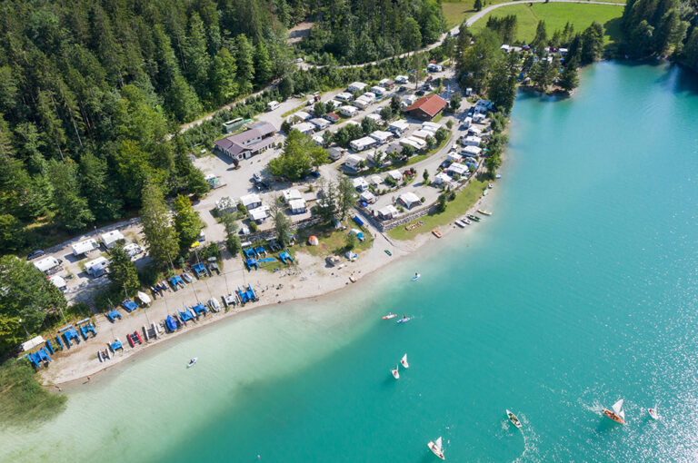 Luftaufnahme eines Campingplatzes am See mit türkisfarbenem Wasser und vielen Segelbooten.